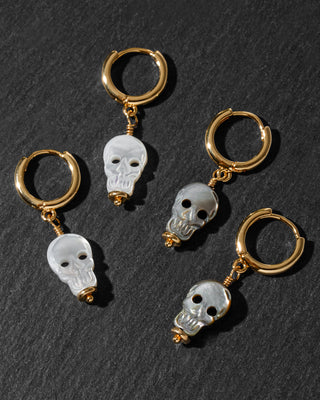 Four gold hoop earrings, each with a small skull charm, arranged on a dark textured slate background.