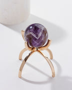 A purple amethyst gemstone sphere on a gold-colored stand, against a white background.