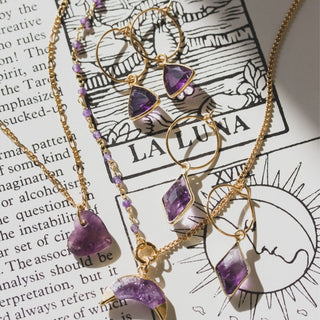 Several amethyst earrings and necklaces laying on a page from an esoteric book with black and white illustration of both the sun and the moon tarot cards.
