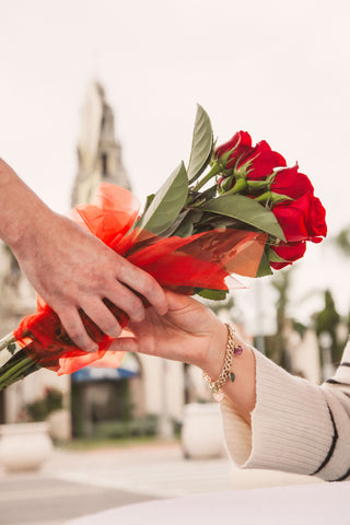Person holding a bouquet of red roses wearing a gemstone heart bracelet with a blurred background