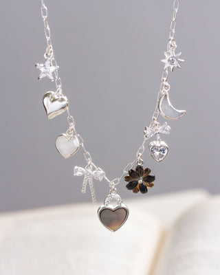 A silver charm necklace featuring various shapes including hearts, a moon, a star, and a flower, with polished silver hearts, CZ pavé bow pendant, and a black lip mother of pearl flower.