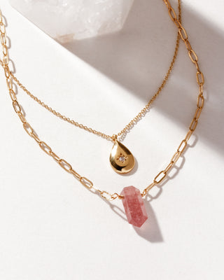 Two layered necklaces, one with a golden teardrop charm and the other with a pink Strawberry Quartz faceted pendant on a white background.