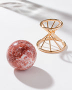 Pink calcite sphere sitting on a white background with a gold stand nearby.