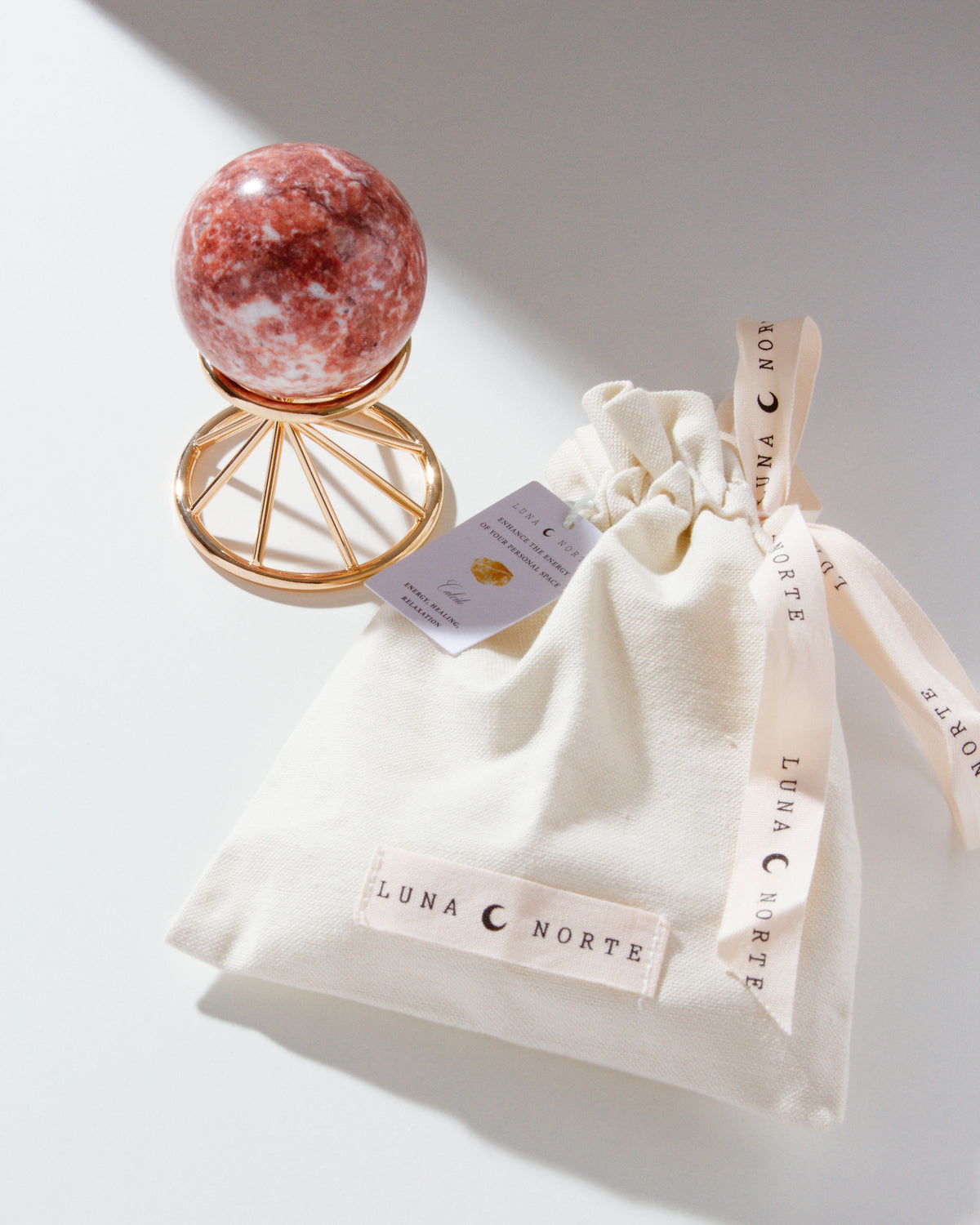 Pink calcite gemstone sphere on gold stand adjacent Luna Norte canvas bag and ribbon packaging.