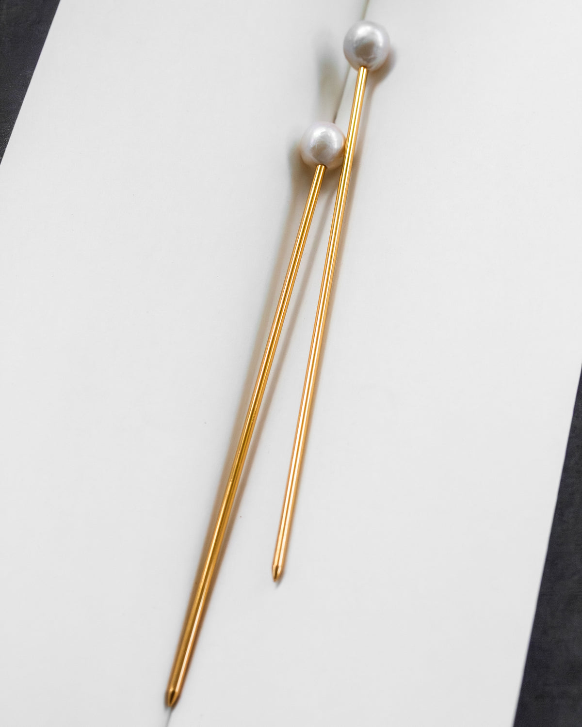Two gold-toned "Ebb and Flow" hair pins, each with a pearl at the top, lying on a white surface.