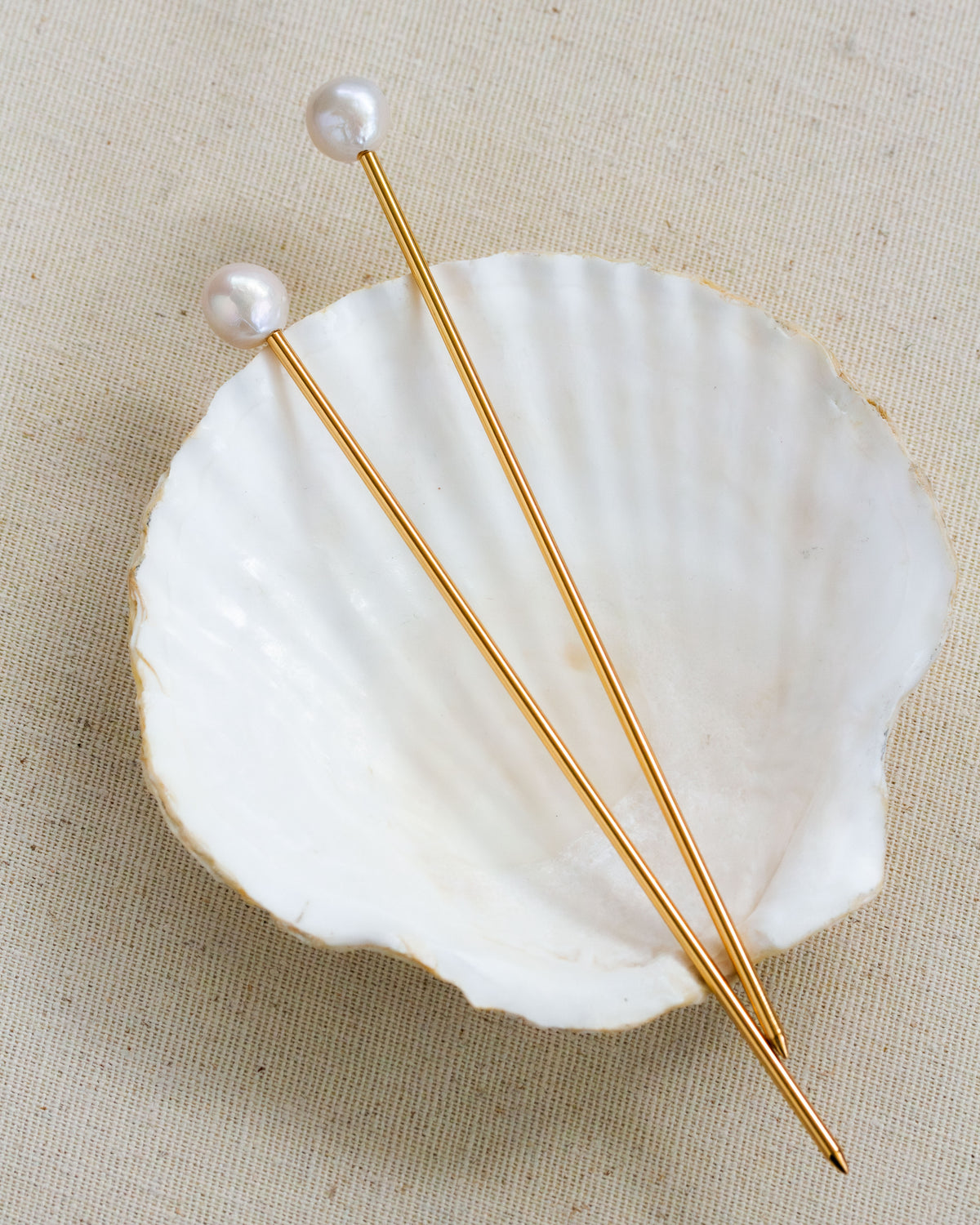 Two Ebb and Flow pearl hair pins with gold stems sit on a white seashell against a tan background.