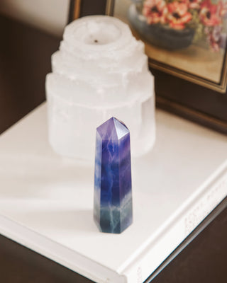 Blue and green flourite obelisk tower gemstone standing on a white background with a painting and candle in the background.