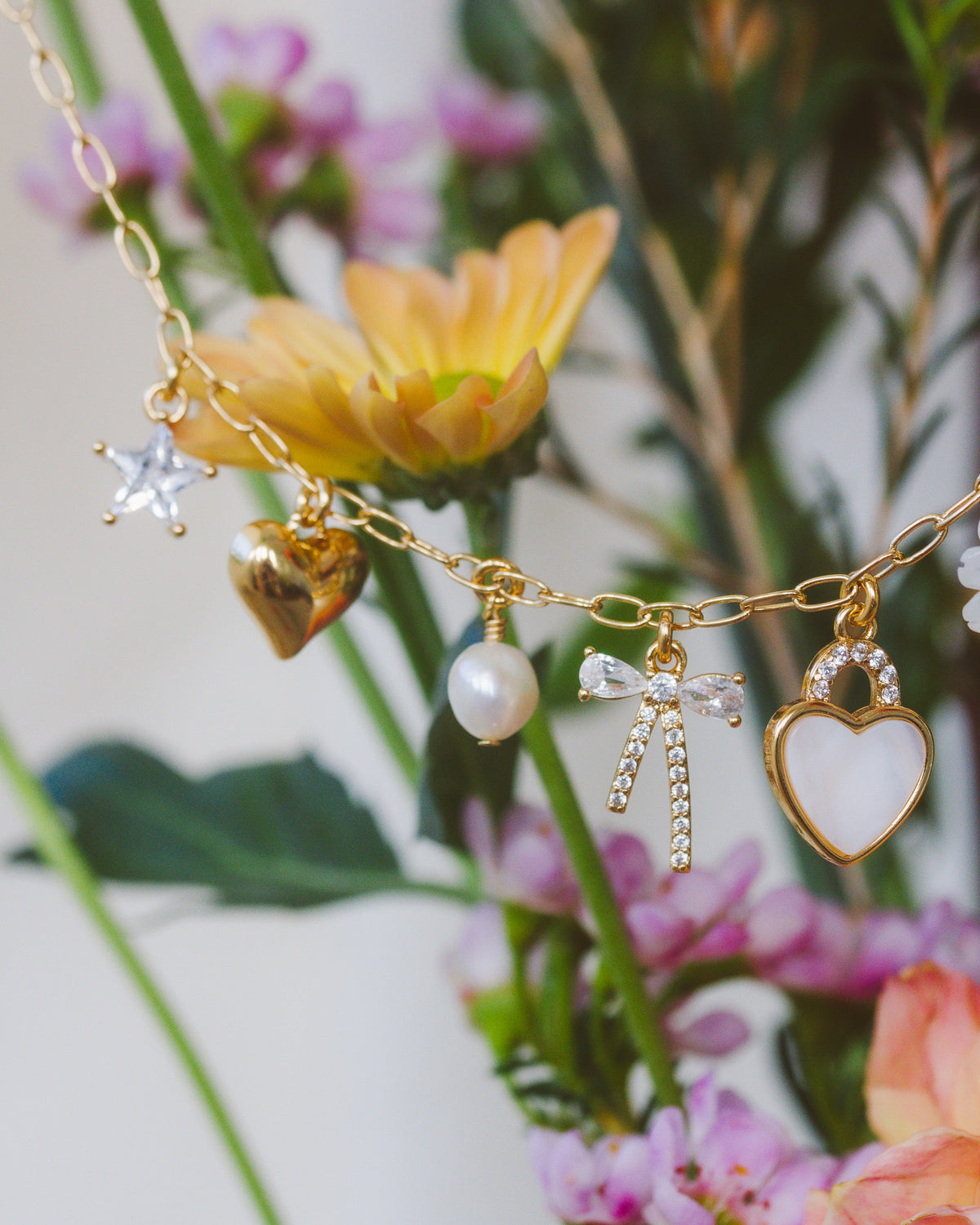 Gold chain necklace with various star, bow, heart and bead charms in front of floral display.