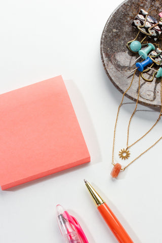Colorful pens and a pink notebook on a white surface with a small round stone dish containing jewelry.