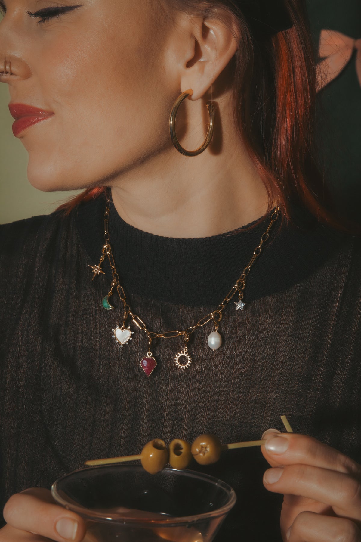 Woman holding a cocktail with olives, wearing a black sweater and gold hoop earrings.