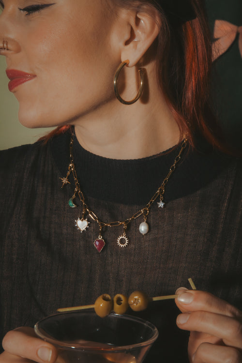 Woman holding a cocktail with olives, wearing a black sweater and gold hoop earrings.