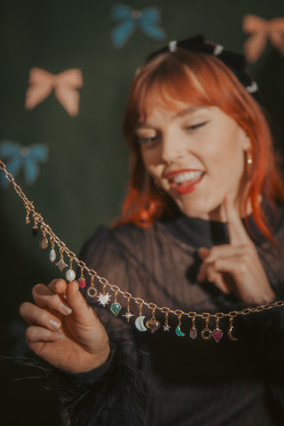 Woman with red hair holding a gold necklace with charms against a dark background with bow decorations.