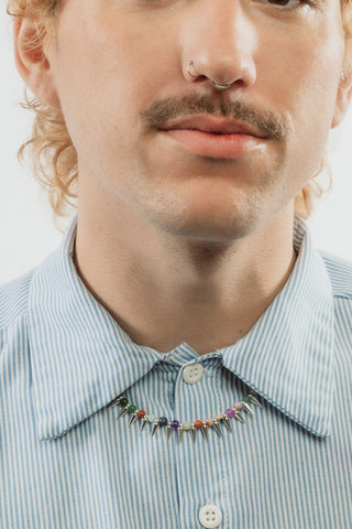 A man with curly light brown hair and a mustache wearing a colorful gemstone spike necklace.