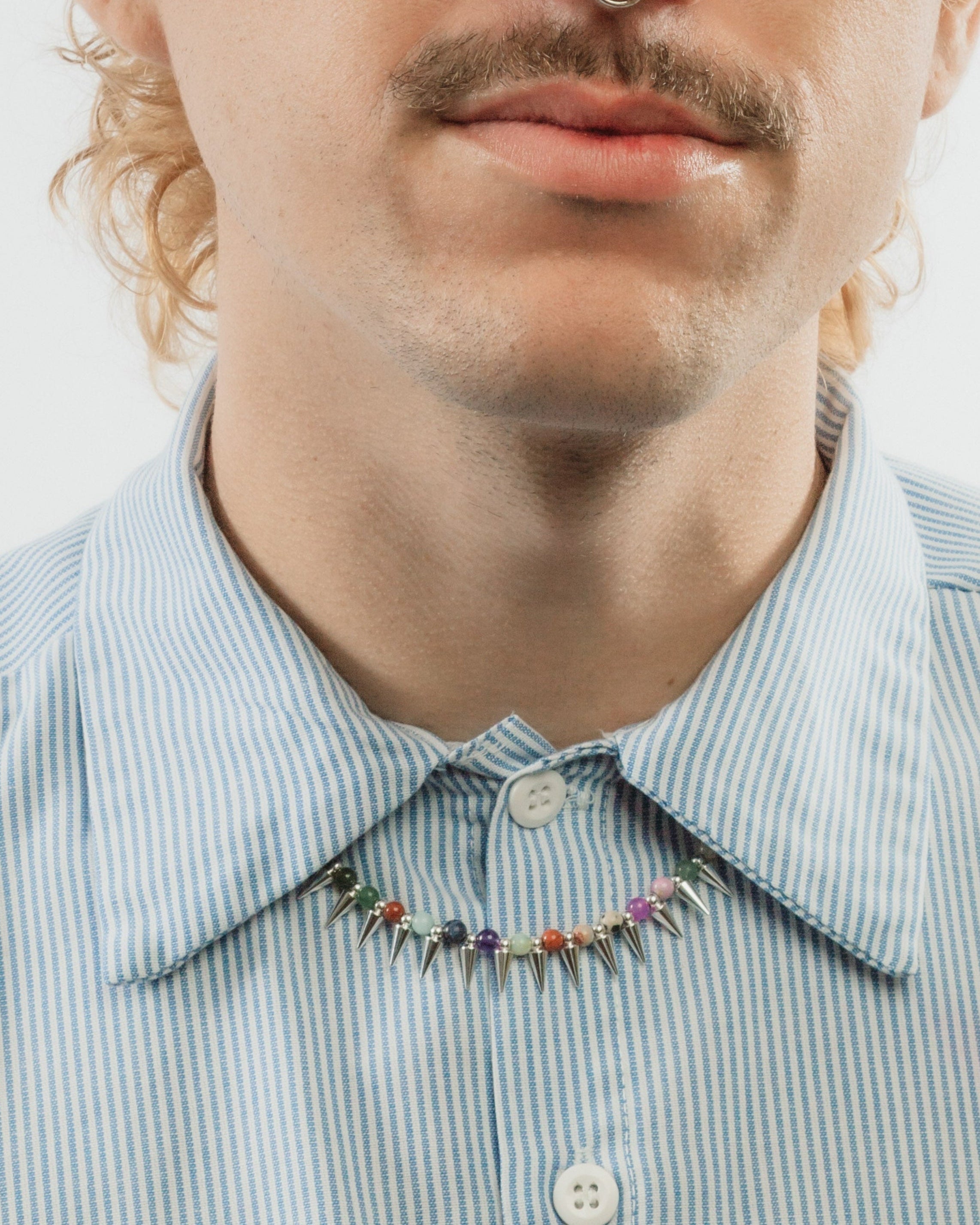 A man with curly light brown hair and a mustache wearing a colorful gemstone spike necklace.