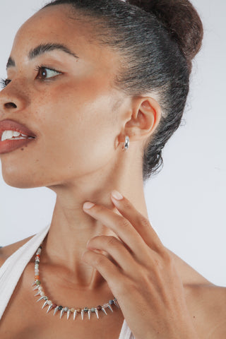 A close-up lifestyle shot of a woman wearing a silver spiked collar necklace and a matching small hoop earring against a light gray background.