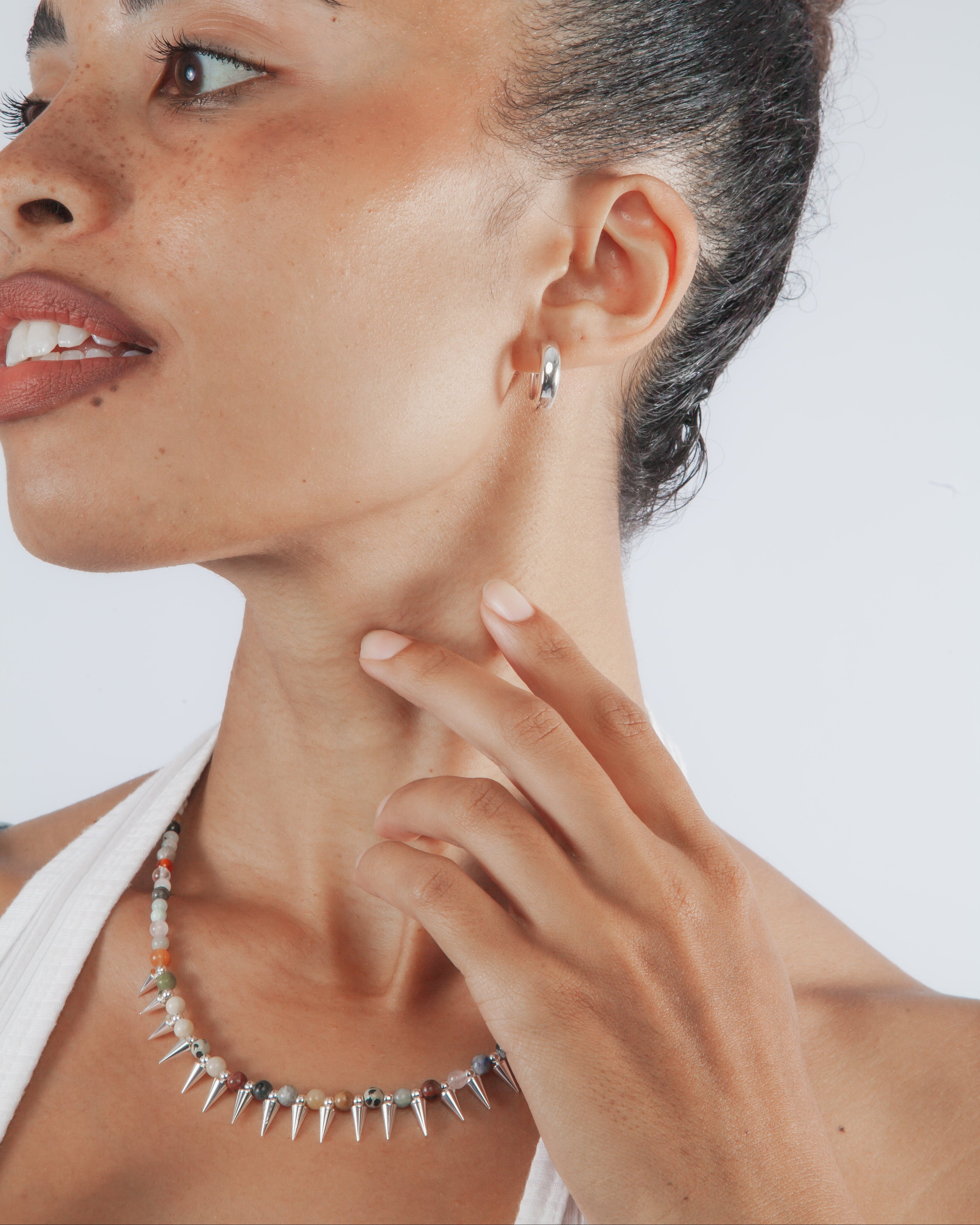 A close-up lifestyle shot of a woman wearing a silver spiked collar necklace and a matching small hoop earring against a light gray background.