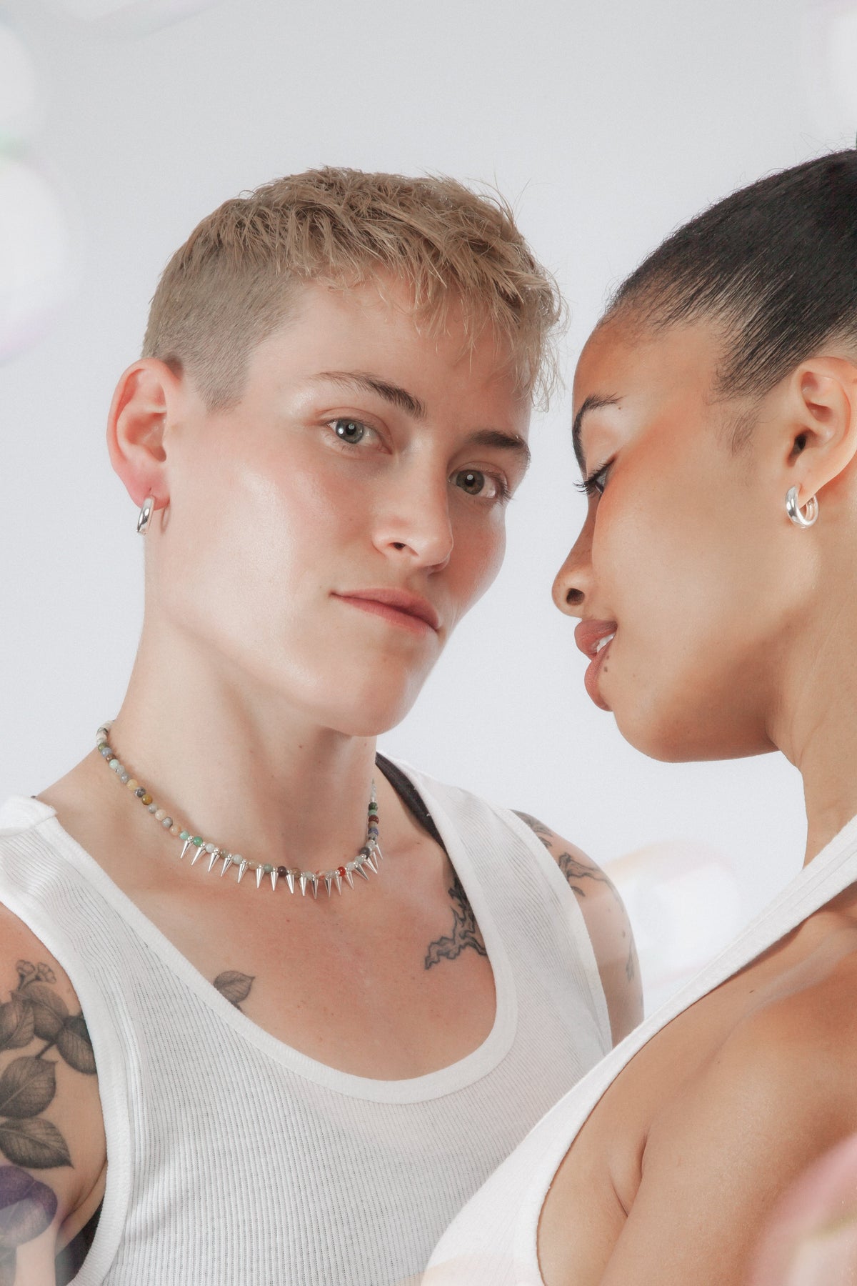 Two people wearing white tank tops with visible tattoos, standing close to each other against a light background wearing gemstone jewelry and silver huggie hoops