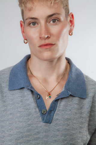 A close-up of a person wearing a blue knit polo collar with a gold necklace featuring a small colorful charm and gold hoop earrings, against a light gray background.