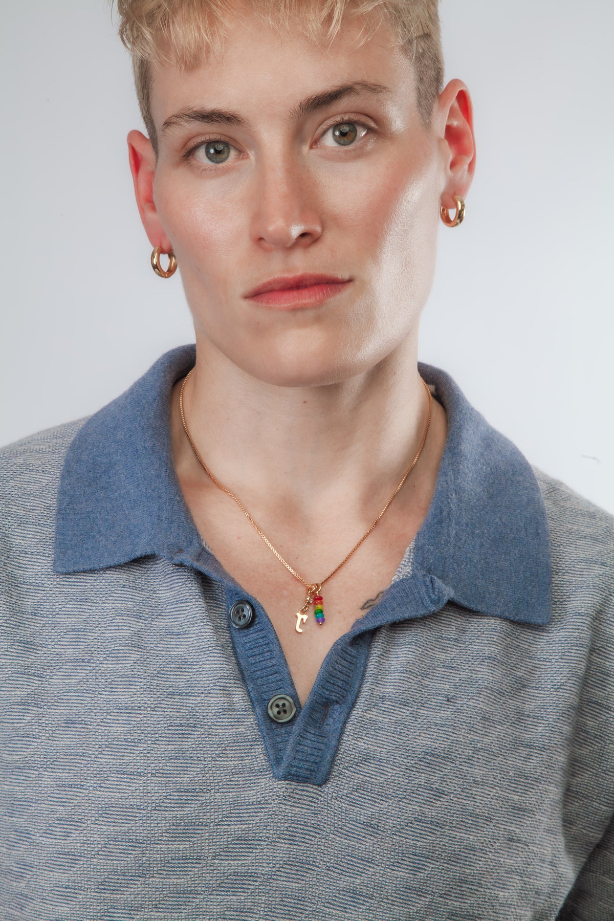 A close-up of a person wearing a blue knit polo collar with a gold necklace featuring a small colorful charm and gold hoop earrings, against a light gray background.