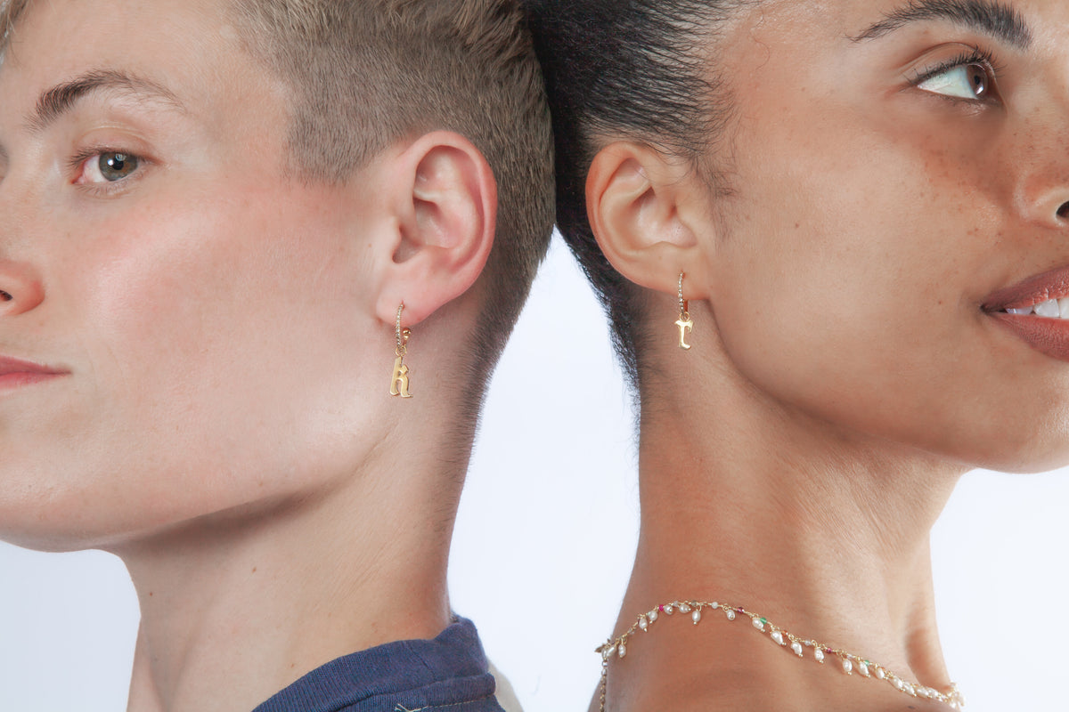 Two people wearing gold initial earrings and a dainty pearl necklace with a plain background