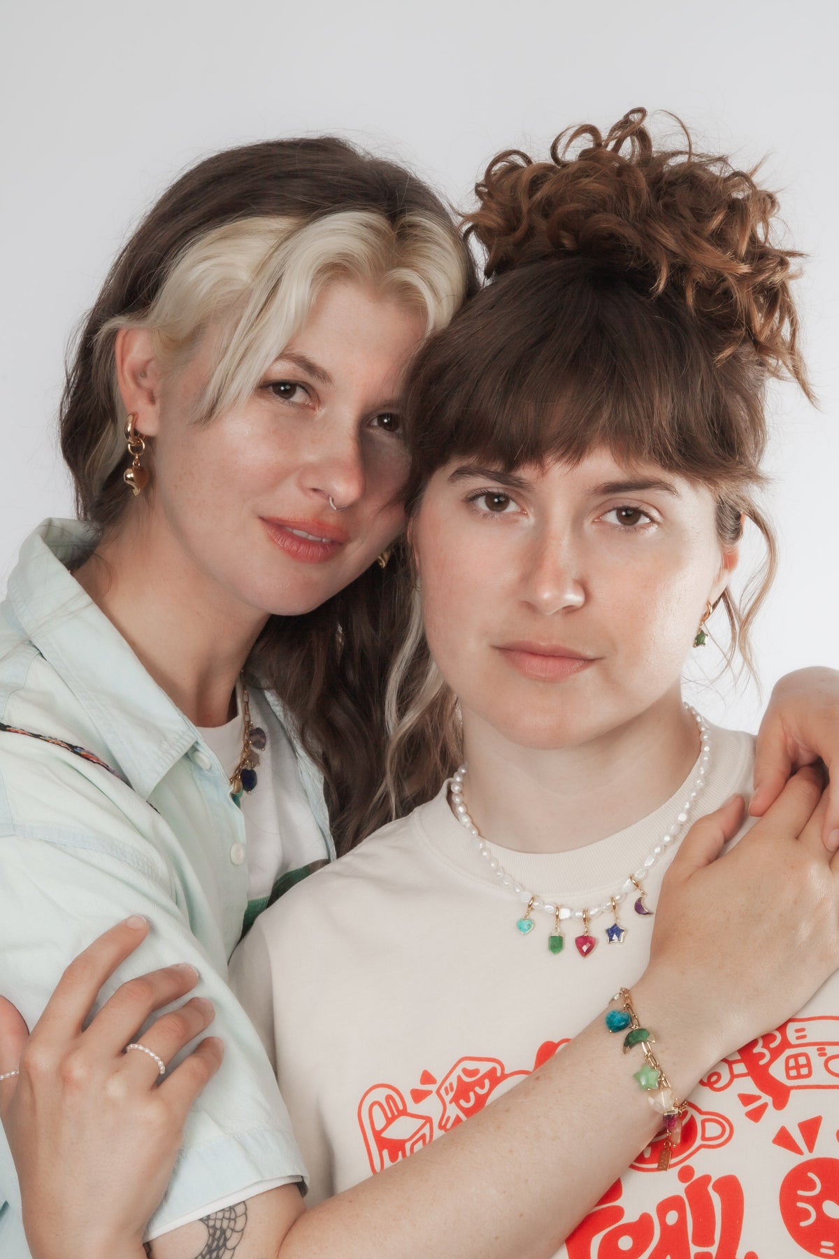 Two people embracing one another with one wearing a white t-shirt with red graphics and the other a denim overshirt. Both wearing various gemstone jewelry styles