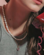 Model holding a red rotary telephone receiver. She is wearing a gold pink heart charm necklace with pearl strand.