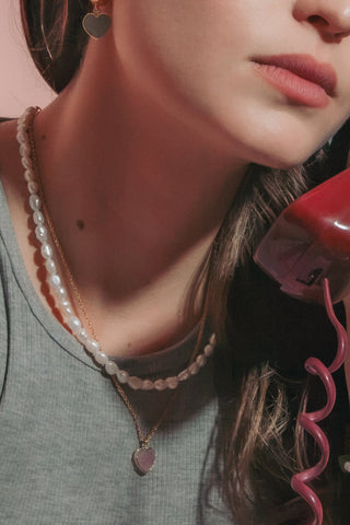 Model holding a red rotary telephone receiver. She is wearing a gold pink heart charm necklace with pearl strand.