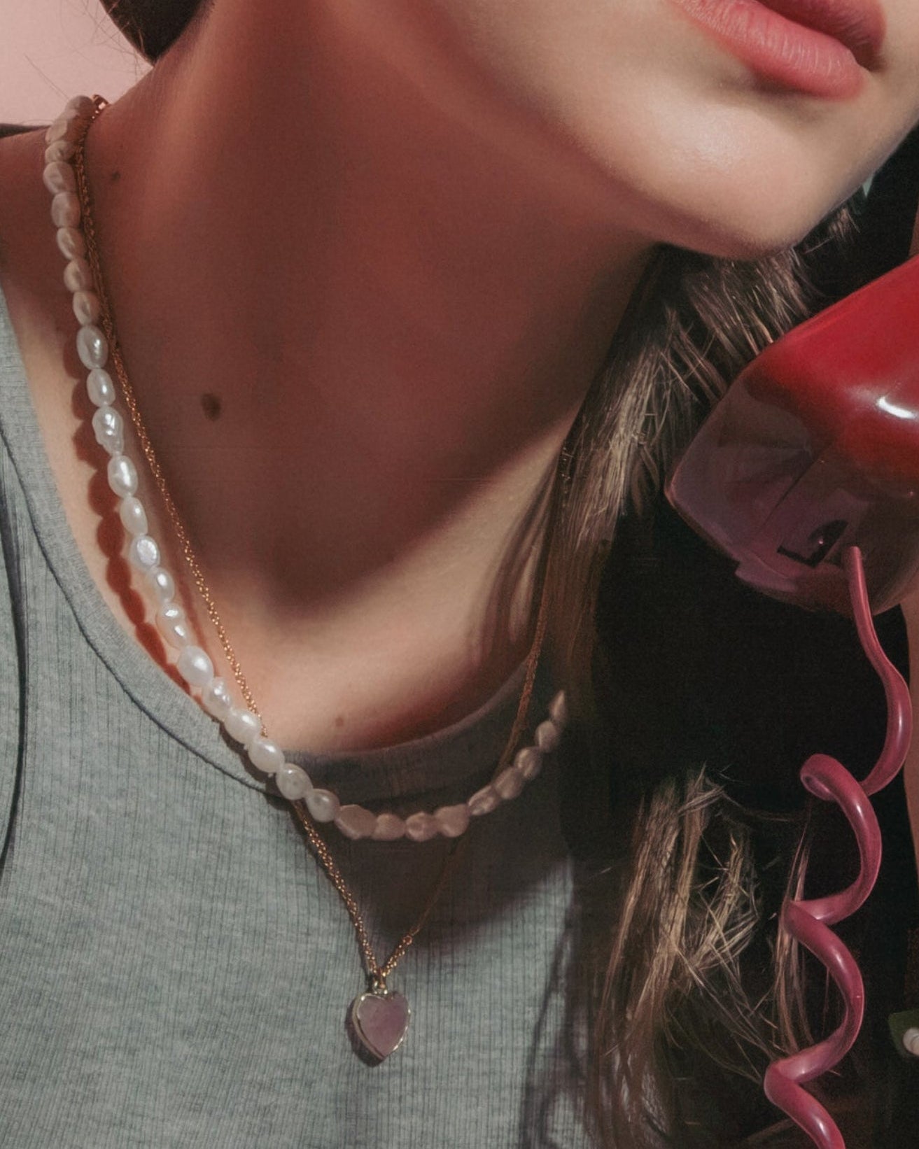 Model holding a red rotary telephone receiver. She is wearing a gold pink heart charm necklace with pearl strand.