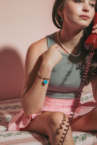 Woman wears layered necklaces and gold charm bracelet with teal heart pendant sits on a bed holding a red phone
