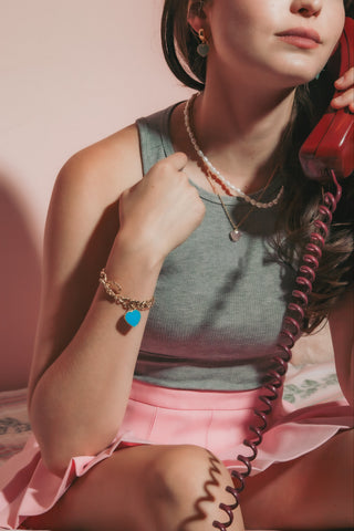 A woman wearing a gold charm bracelet with a blue heart pendant and layered necklaces sits on a bed while holding a red rotary telephone receiver. The product featured is the gold bracelet with the blue heart charm.