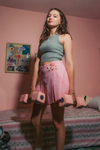 Woman in a gray crop top and pink mini skirt with gemstone pins holds pink dumbbells in pink bedroom