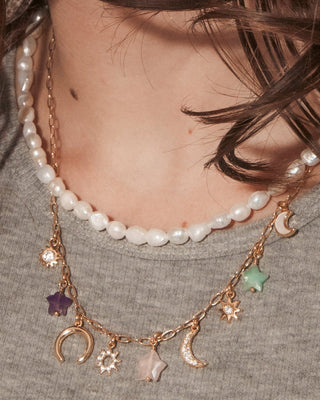 A close-up portrait of a woman with long dark brown hair wearing layered necklaces, including a pearl necklace and a gold-toned charm necklace with crescent moon and star details.