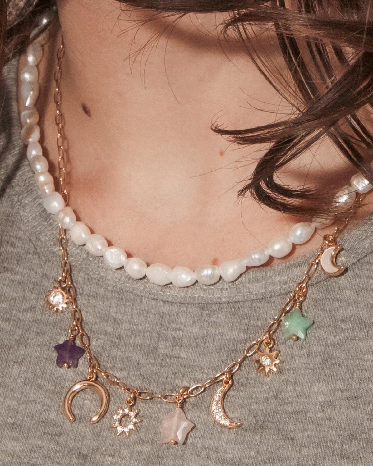 A close-up portrait of a woman with long dark brown hair wearing layered necklaces, including a pearl necklace and a gold-toned charm necklace with crescent moon and star details.