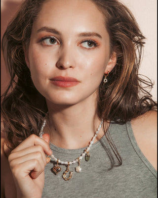 A woman with wavy brown hair wearing a pearl beaded necklace with various charms and matching sun charm drop earrings.