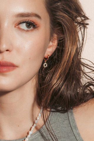 A woman with wavy brown hair wearing gold clear CZ sun charm huggie hoop earrings.