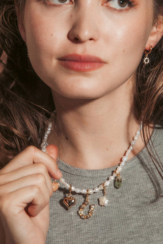 A woman with wavy brown hair wearing a pearl beaded necklace with gold charms and matching sun charm drop earrings.