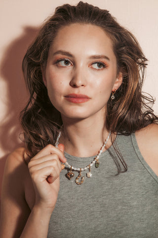 A woman with wavy brown hair wearing a pearl beaded necklace with small charms and matching gold charm earrings, posed against a warm pink background.