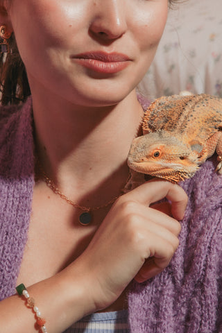 A woman wearing a purple knit cardigan and a striped top holds an orange bearded dragon on her shoulder wearing a round gemstone pendant.