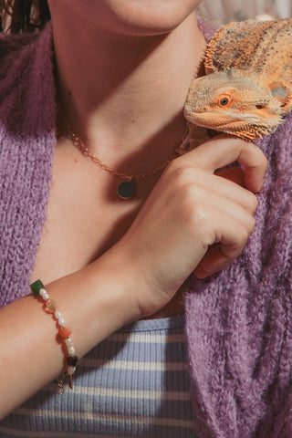 A woman wearing a purple knit cardigan and a striped top holds an orange bearded dragon wearing a beaded pearl gemstone bracelet.