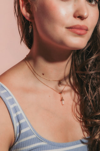 A close-up portrait of a young woman with long, wavy brown hair, wearing a light blue and white striped tank top and layered gold necklaces.