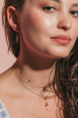 A close-up portrait of a young woman with long, wavy brown hair, wearing a light blue and white striped tank top and layered gold necklaces, set against a solid dusty pink background.