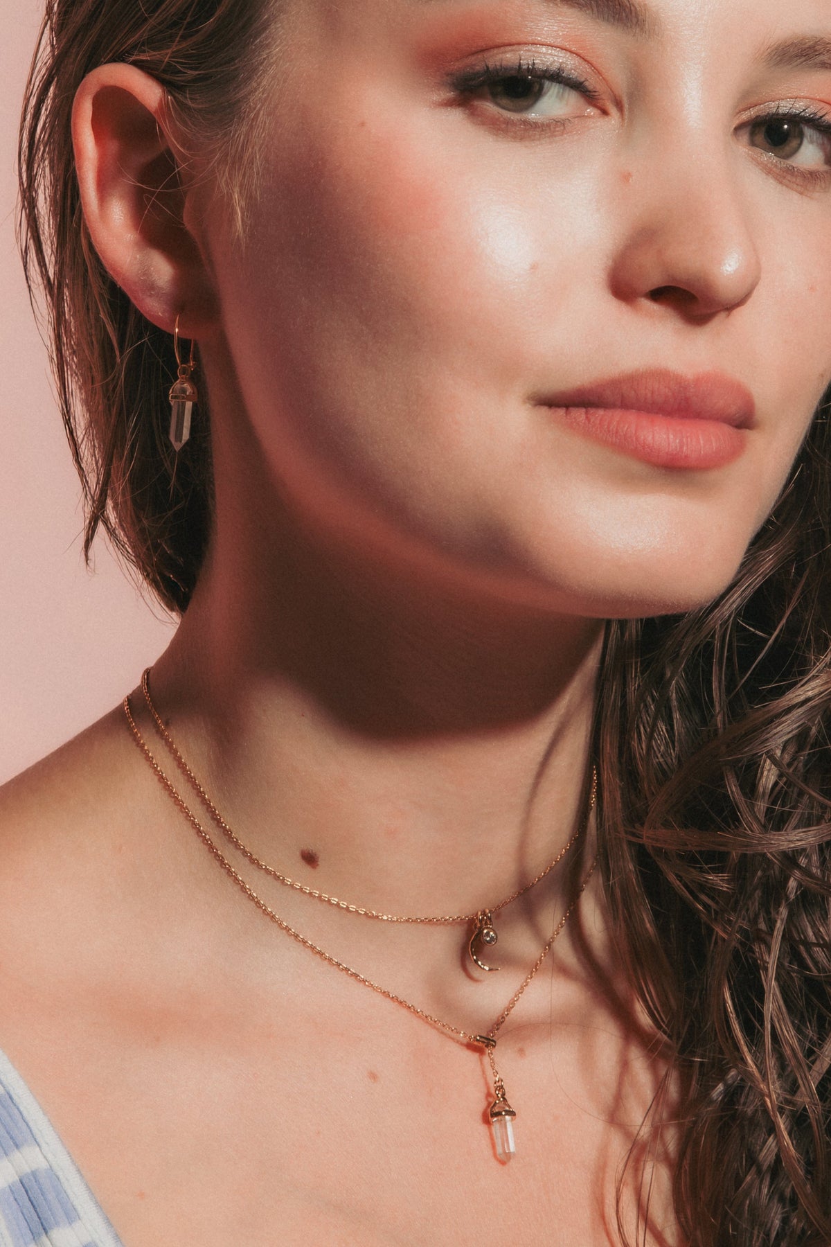 A close-up portrait of a young woman with long, wavy brown hair, wearing a light blue and white striped tank top and layered gold necklaces, set against a solid dusty pink background.