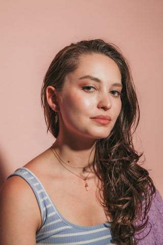 Young woman with wavy brown hair, wearing striped tank top and layered gold gemstone necklaces