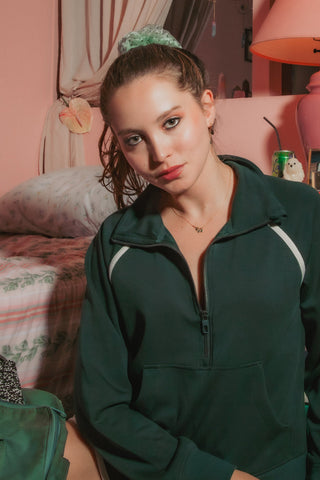 A woman in a dark green half-zip sweatshirt with white shoulder stripes poses in a pink bedroom