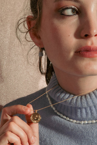 A woman wearing a light blue ribbed turtleneck sweater and a pearl necklace, holding a gold moon detail  pendant necklace. 