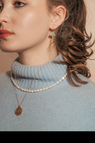 A woman in a light blue turtleneck sweater wearing a beaded white pearl necklace and a gold moon pendant necklace, with a gemstone cloud earring.