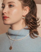 A woman in a light blue turtleneck sweater wearing a delicate white pearl necklace and a gold pendant necklace, with a green cloud  earring.