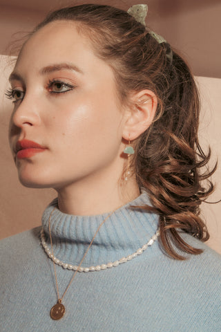 A woman in a light blue turtleneck sweater wearing a delicate white pearl necklace and a gold pendant necklace, with a green cloud  earring.