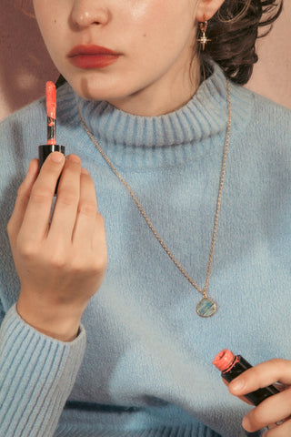 A close-up lifestyle shot of a woman wearing a light blue knit turtleneck sweater, holding a tube of coral-pink lipstick in both hands wearing a longer length necklace.