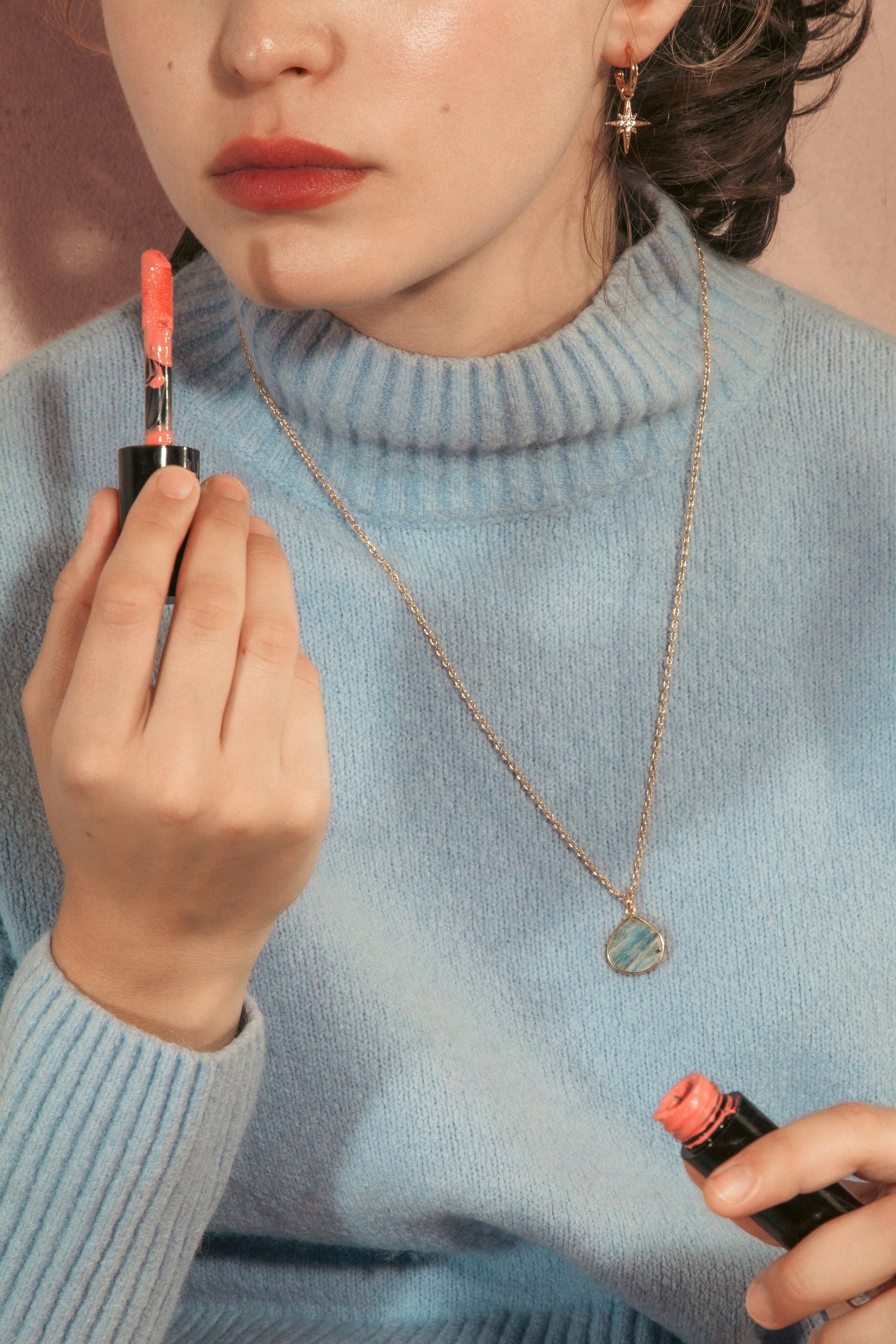 A close-up lifestyle shot of a woman wearing a light blue knit turtleneck sweater, holding a tube of coral-pink lipstick in both hands wearing a longer length necklace.